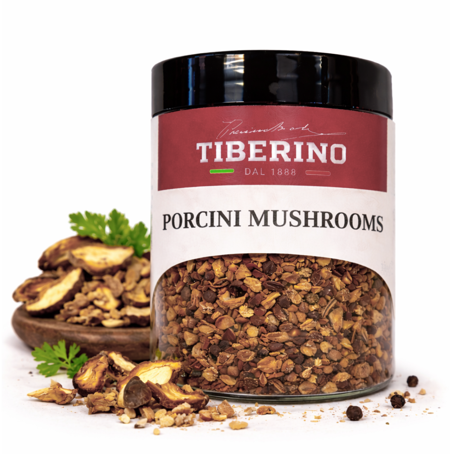 Barattolo di vetro trasparente con coperchio nero, etichetta rossa Tiberino 1888 e Porcini Mushrooms, contenente funghi porcini secchi in briciole marroni. Accanto, una ciotola di legno con funghi secchi e prezzemolo fresco.