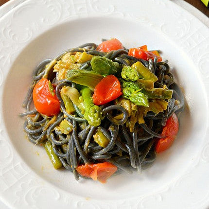 PIATTO BIANCO SU TAVOLA DI LEGNO DI SPAGHETTI NERI CON PUNTE DI ASPARAGO VERDE E POMODORO 