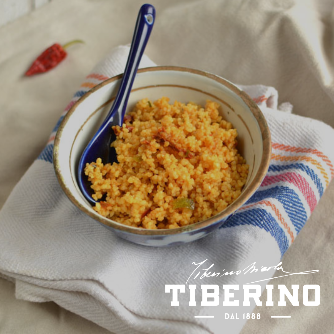 Couscous alla Marocchina in ciotola bianca e blu con cucchiaio blu, su strofinaccio a righe rosse e blu, peperoncino rosso sullo sfondo.