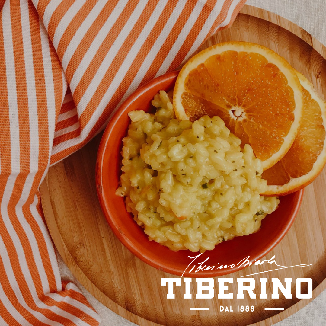 Risotto giallo cremoso in ciotola arancione, guarnito con due fette d'arancia su vassoio di legno, con tovagliolo a righe bianche e arancioni. Logo Tiberino 1888.