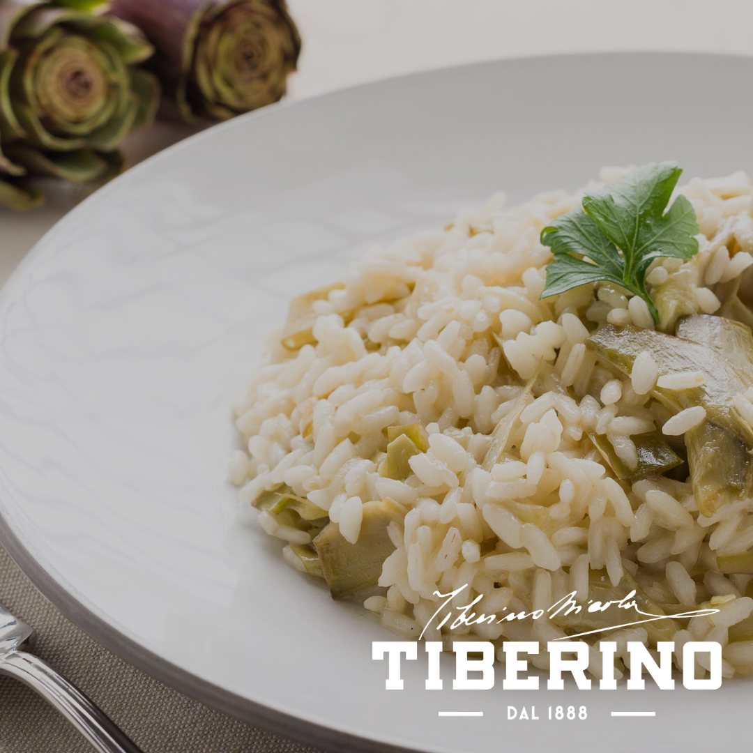 Risotto con carciofi alla Romana in un piatto bianco, guarnito con prezzemolo, con carciofi freschi sullo sfondo.
