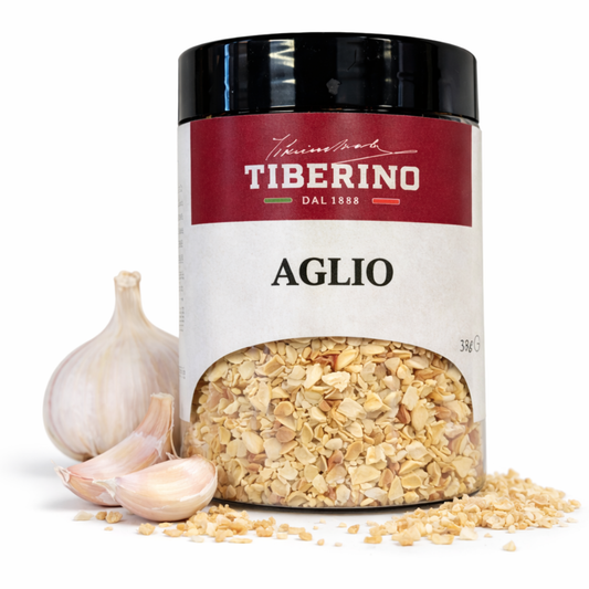 Barattolo trasparente Tiberino 1888 di aglio essiccato in granuli da 38g con etichetta rossa e bianca, accanto a bulbo e spicchi d'aglio freschi.