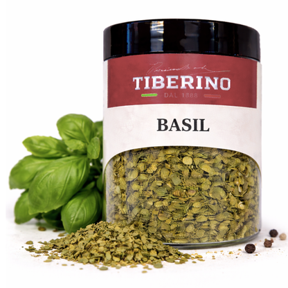 Vasetto di vetro trasparente Tiberino 1888 con basilico liofilizzato verde chiaro, etichetta rossa e nera, accanto a foglie di basilico fresco e grani di pepe.