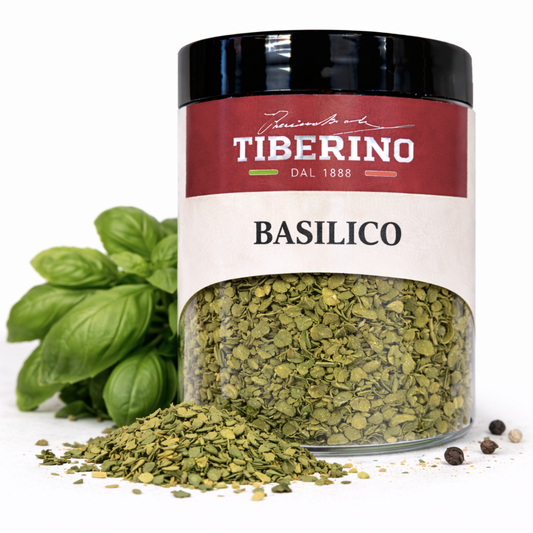 Barattolo in vetro trasparente di Basilico Liofilizzato Tiberino 1888 con etichetta rossa e beige, accanto a foglie fresche di basilico verde e grani di pepe.