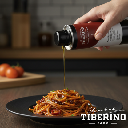 Mano versa Condimento al Peperoncino Tiberino 1888 da lattina scura da 250ml su spaghetti rossi in piatto nero.