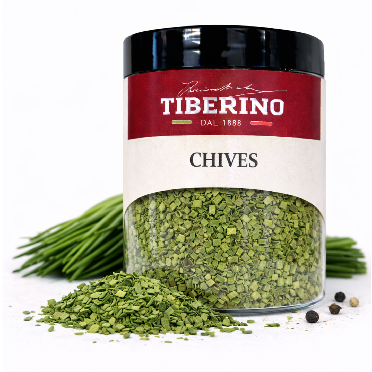 Barattolo trasparente Tiberino 1888 con erba cipollina essiccata, etichetta rossa e bianca, accanto a erba cipollina fresca e grani di pepe.
