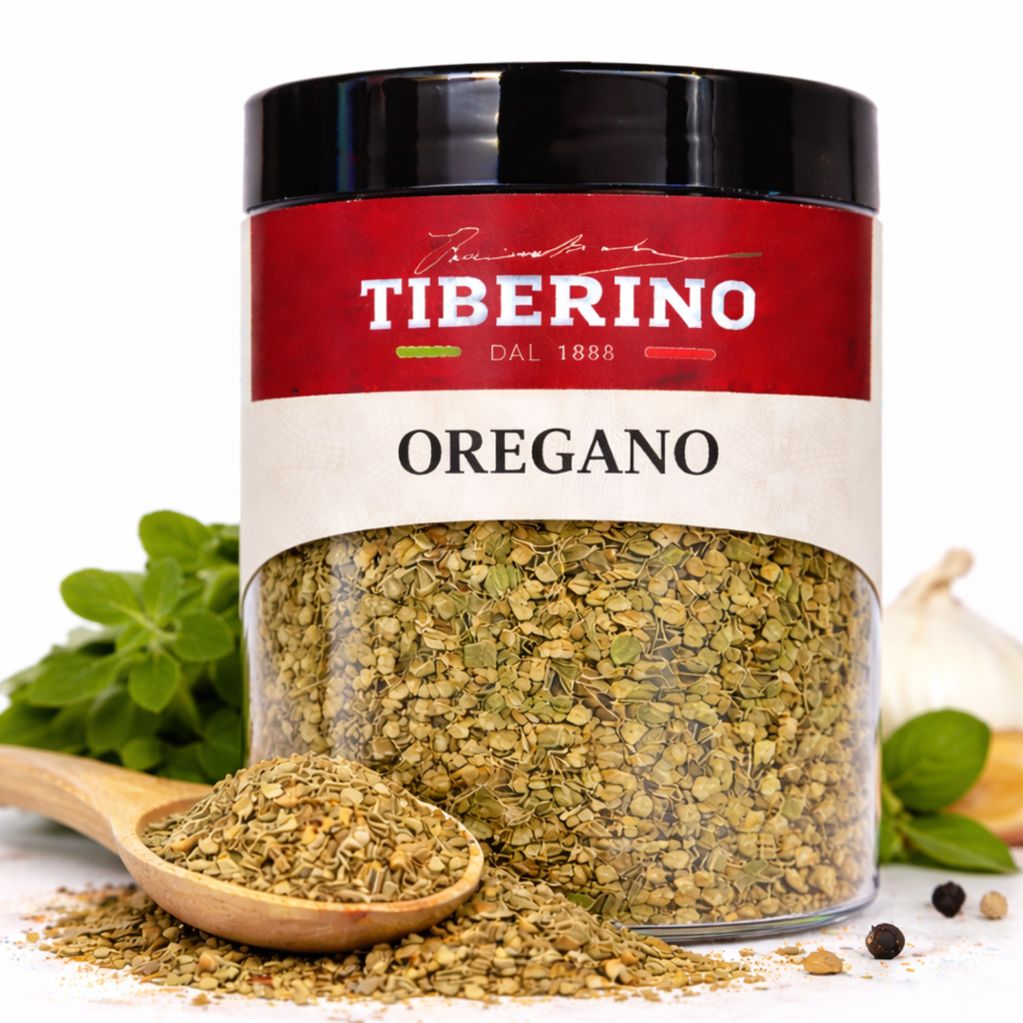Barattolo trasparente di origano essiccato Tiberino 1888 con etichetta rossa, cucchiaio di legno e aglio.