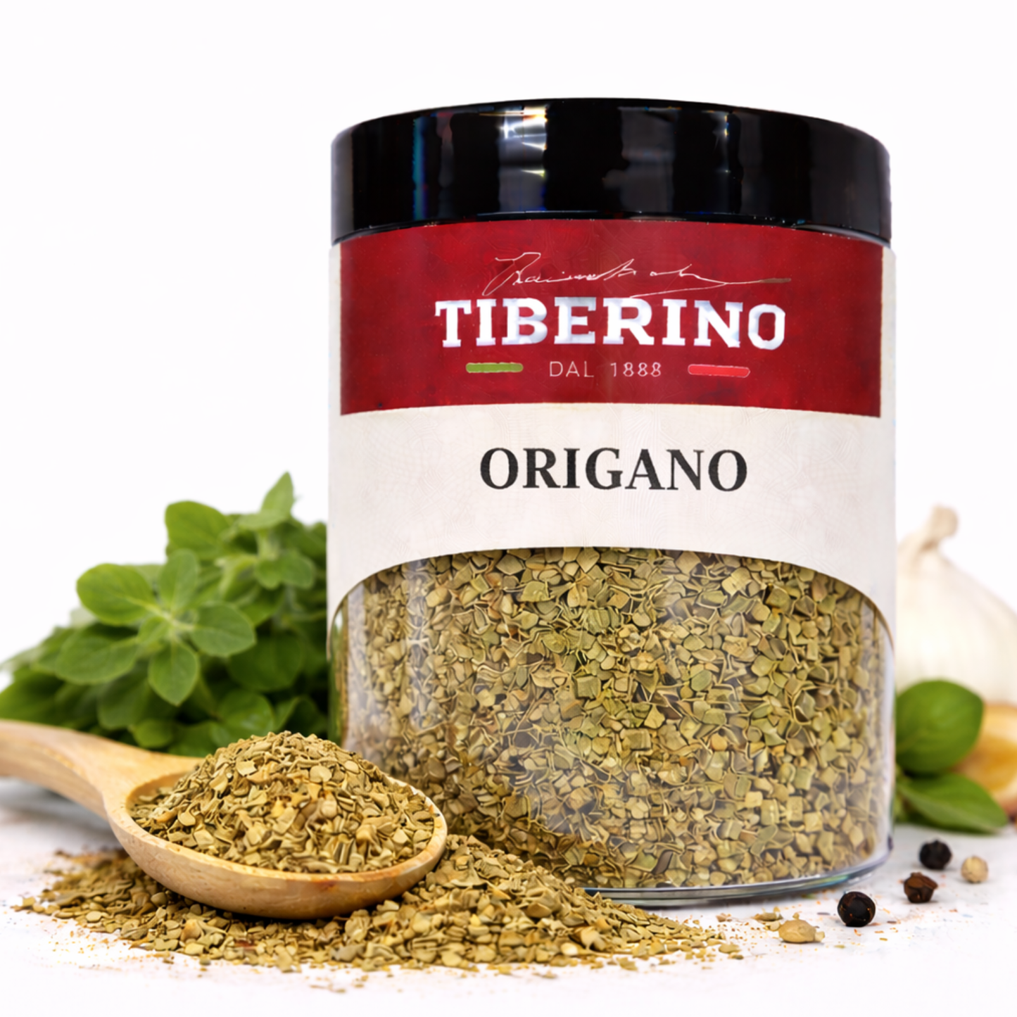 Barattolo trasparente di origano essiccato Tiberino 1888 con etichetta rossa e beige, cucchiaio di legno, foglie fresche e aglio.