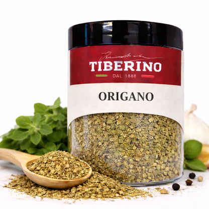 Barattolo trasparente di origano essiccato Tiberino 1888 con etichetta rossa e beige, cucchiaio di legno, foglie fresche e aglio.
