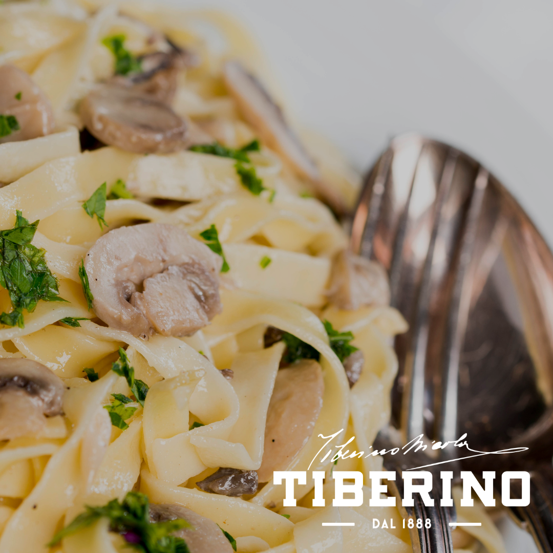 Pappardelle all'uovo con funghi porcini e prezzemolo fresco, servite in un piatto bianco con posate d'argento, logo Tiberino 1888.
