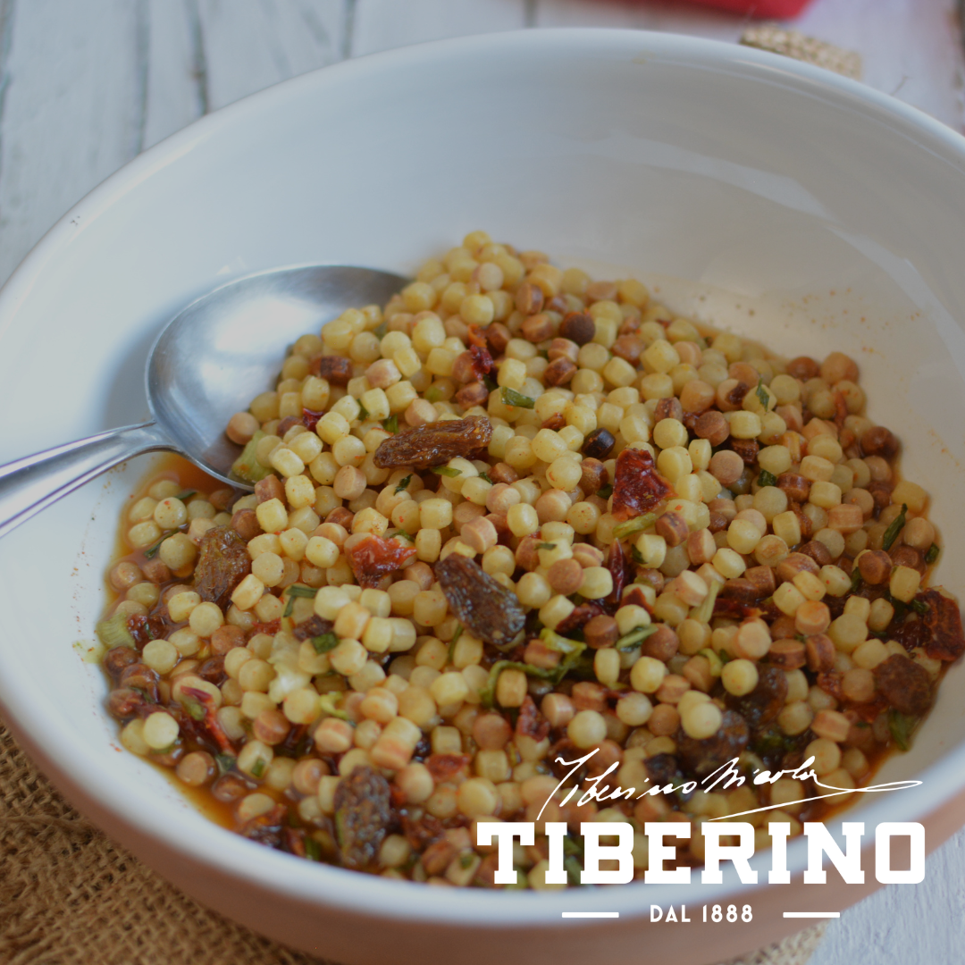 Pasta Fregola Sarda ai peperoni rossi e capperi al sale in una ciotola bianca con cucchiaio d'argento.