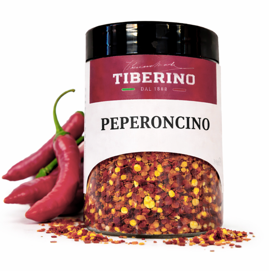 Barattolo trasparente di Peperoncino Frantumato Tiberino 1888 da 30g, con etichetta rossa e nera, affiancato da peperoncini rossi freschi.