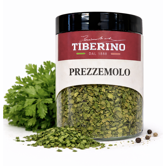 Barattolo di vetro trasparente Tiberino 1888 con prezzemolo secco verde, tappo nero, etichetta rossa e beige, accanto a un mazzetto di prezzemolo fresco e grani di pepe.