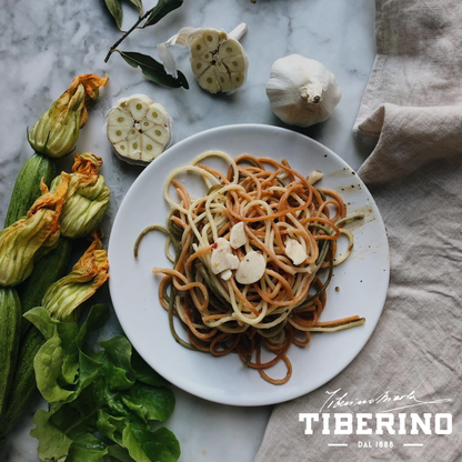 Spaghetti tricolore aglio, olio e peperoncino su piatto bianco, con aglio, zucchine, fiori di zucca e basilico su marmo.