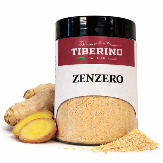 Barattolo di vetro trasparente con coperchio nero e etichetta rossa Tiberino 1888, contenente zenzero in polvere beige chiaro, affiancato da radici di zenzero fresco.